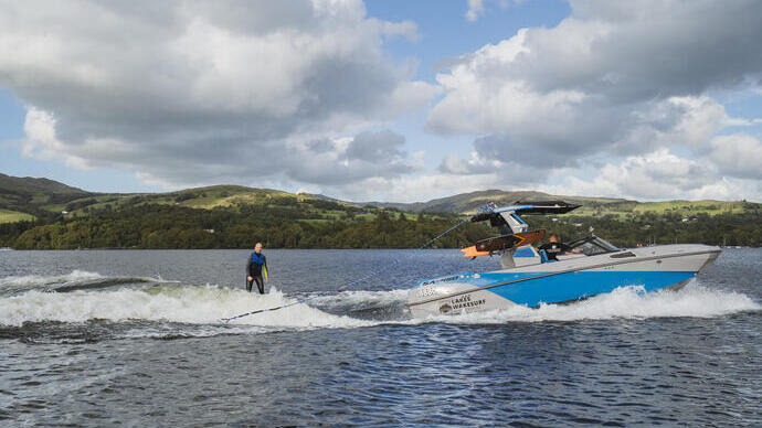 Person wakeboarding behind a blue speedboat on a lake with green hills and cloudy sky in the background.