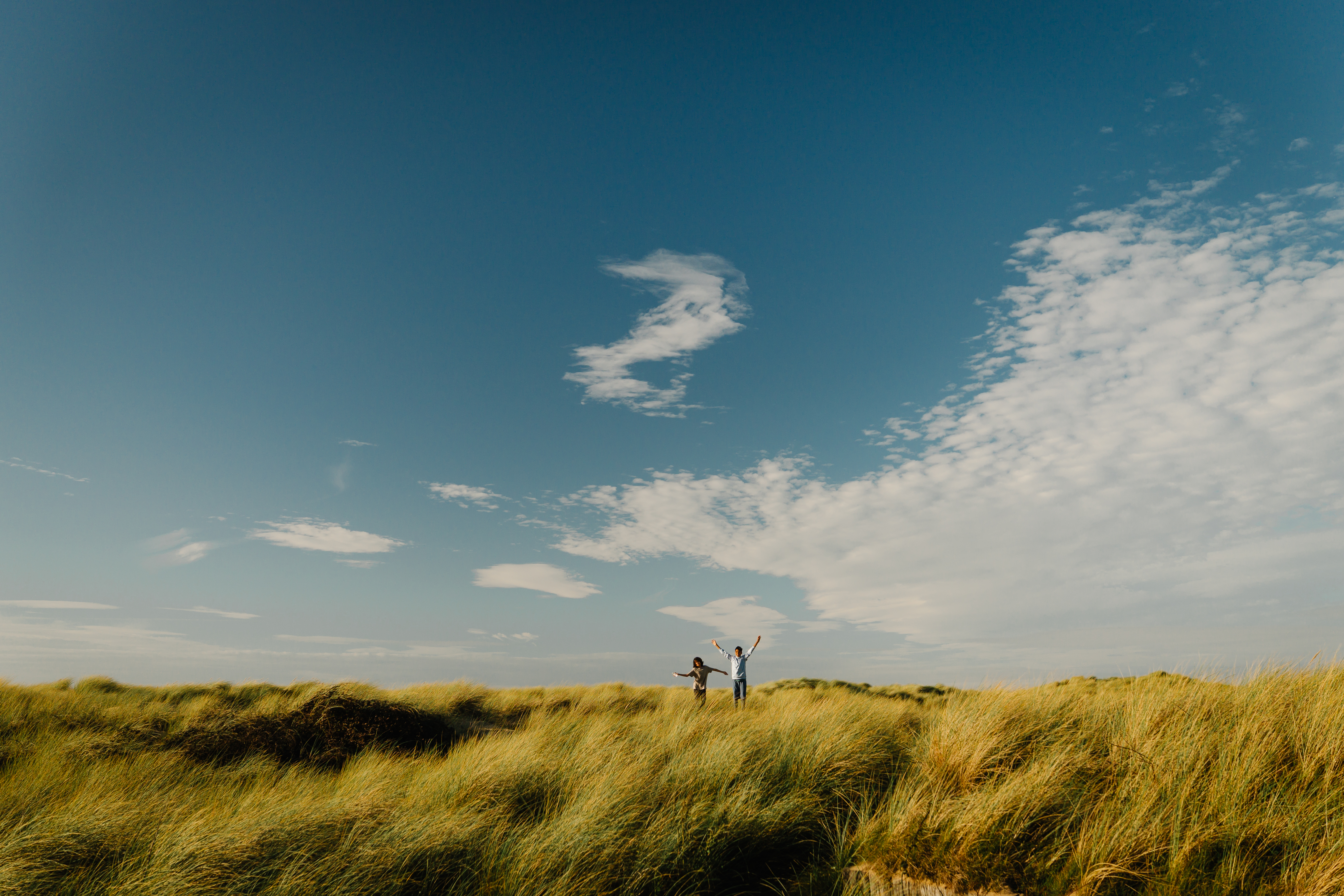 Two people enjoying tall grass dunes under a vast blue sky with scattered clouds, evoking freedom and nature.