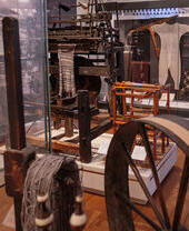 Museum display featuring historical textile machinery, spinning wheel, yarn, and vintage garment in a glass case.