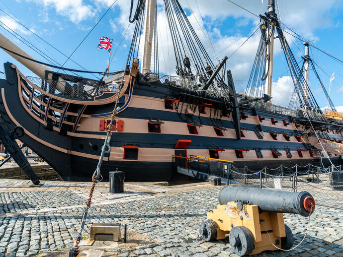Historic wooden warship docked on cobblestone with a cannon in the foreground, under a blue sky with some clouds.