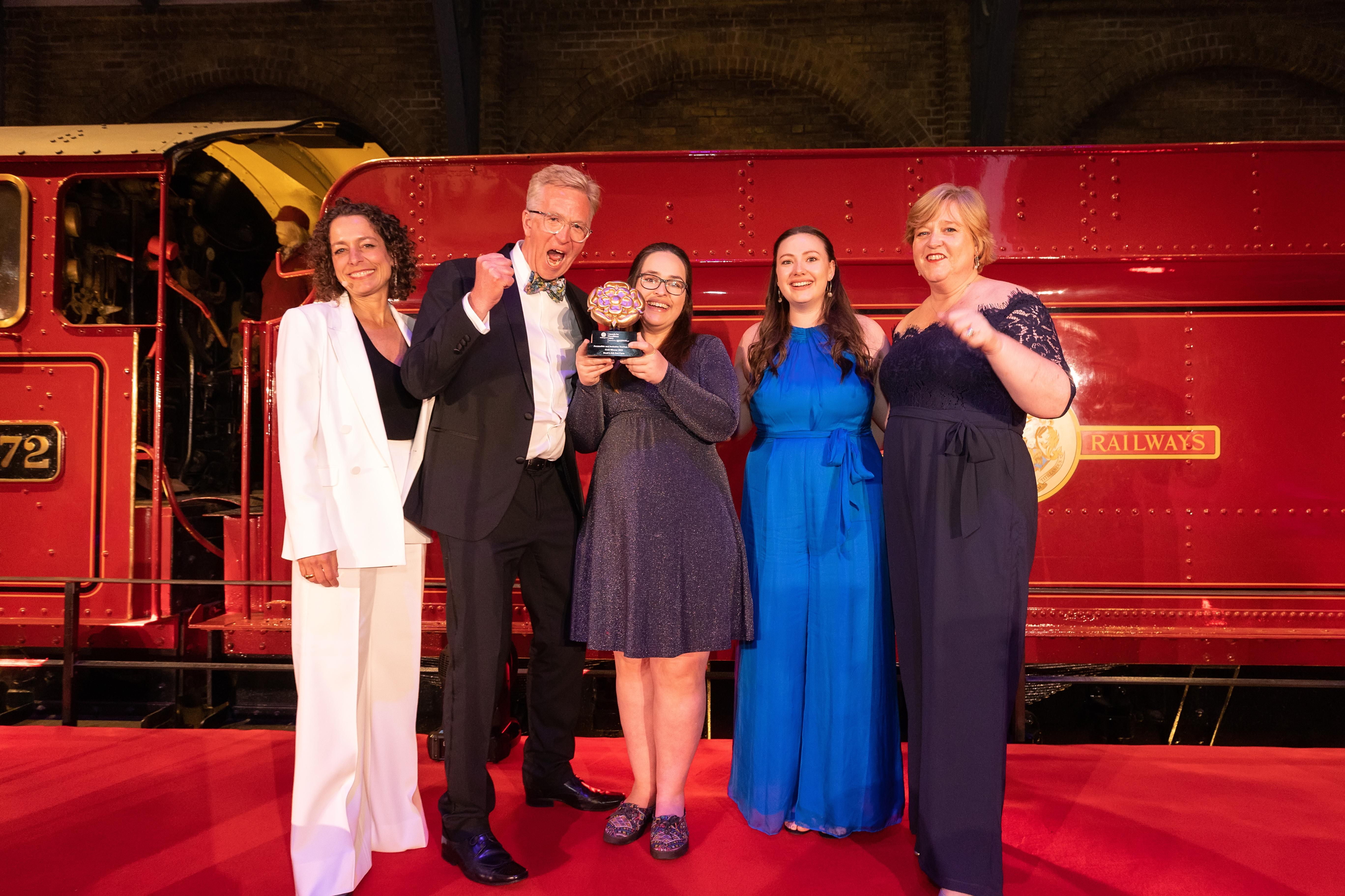Photo of Gold Award Winners for the Accessible & Inclusive Tourism Award at the VisitEngland Awards for Excellence 2023 - Noah's Ark Zoo Farm (Representatives from Noah's Ark Zoo Farm, VisitEngland Director of HR and Professional Services Debra Lang, Relationship Director for Hospitality & Leisure at Corporate Banking Barclays Andy Mayne, Compere Alex Polizzi) - in front of the Hogwarts Express on stage