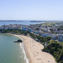 An overhead shot of a sandy beach lined with sunbathers, surrounded by brightly coloured townhouses and countryside.