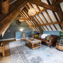 Lounge area at the top of a timber framed building, with exposed ceiling beams, a large leather sofa and wooden coffee table facing a flat-screen TV