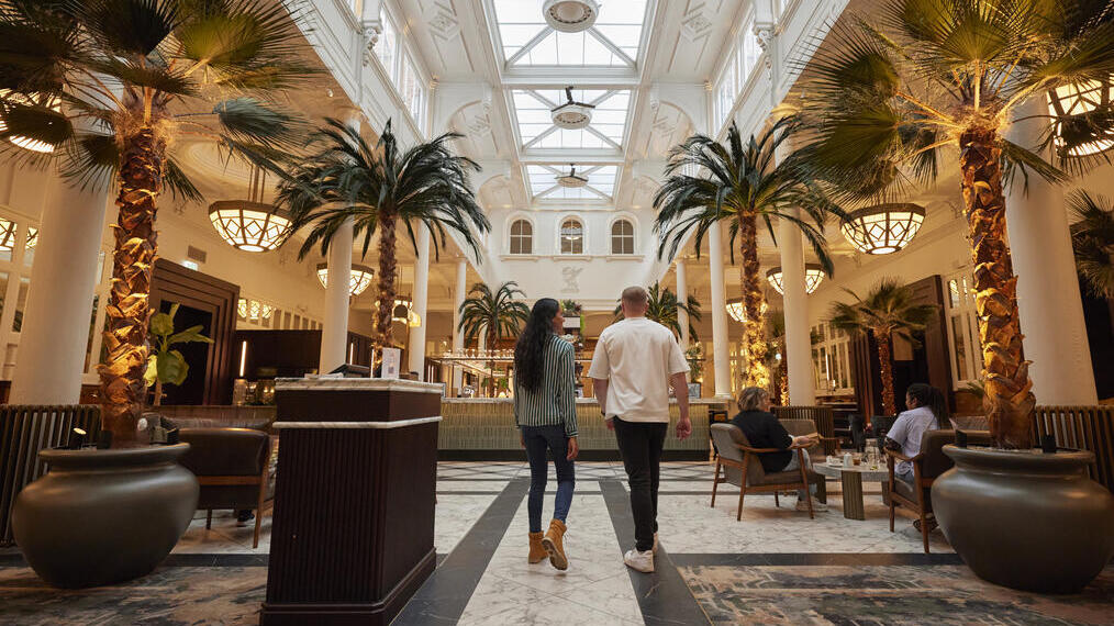 Elegant indoor lounge with palm trees, marble floors, skylight ceiling, and people relaxing or walking through the space.