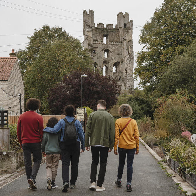 Five people walk towards a stone tower ruin, with a house and garden beside the road. Trees and greenery surround the area.