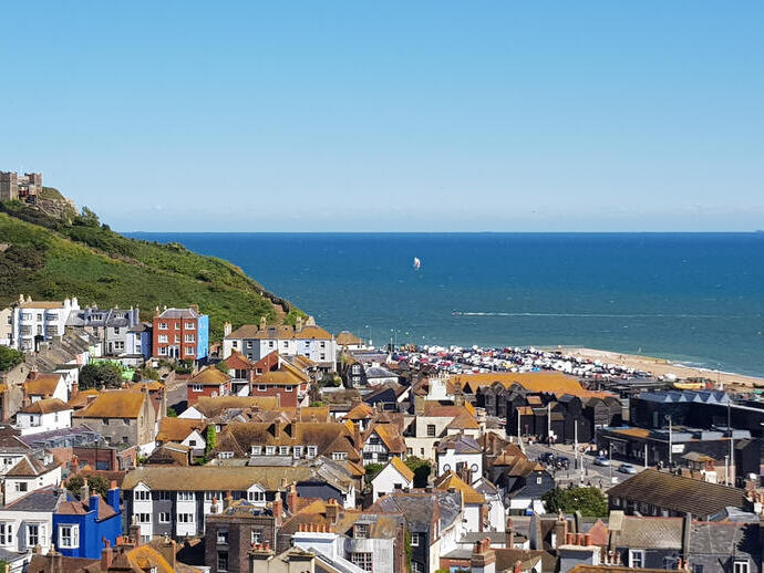 Coastal town with clustered houses, a green hill with a castle, and a beach by the blue sea under a clear sky.