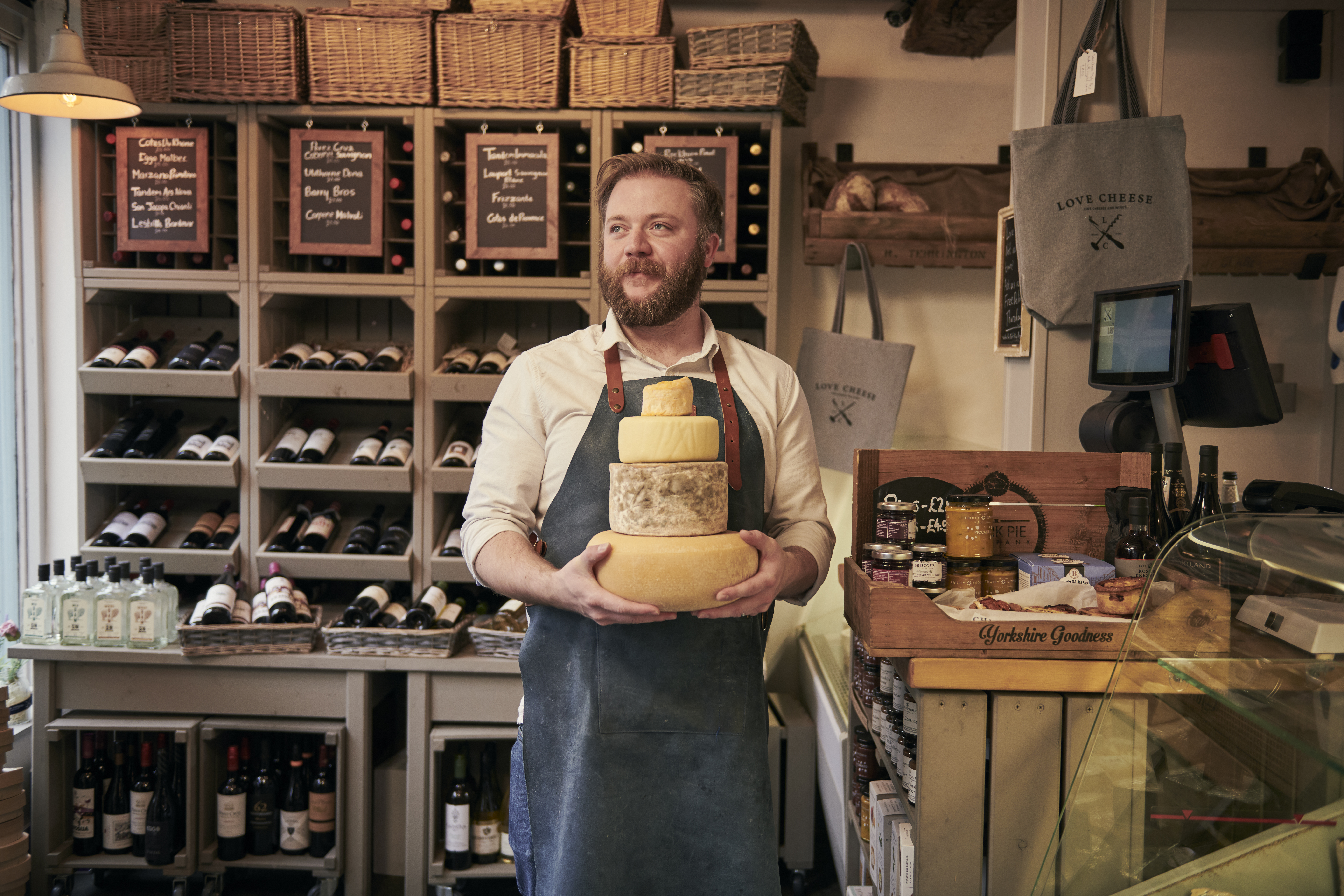 Man wearing blue apron holding a stack of cheeses in a cheese and wine shop