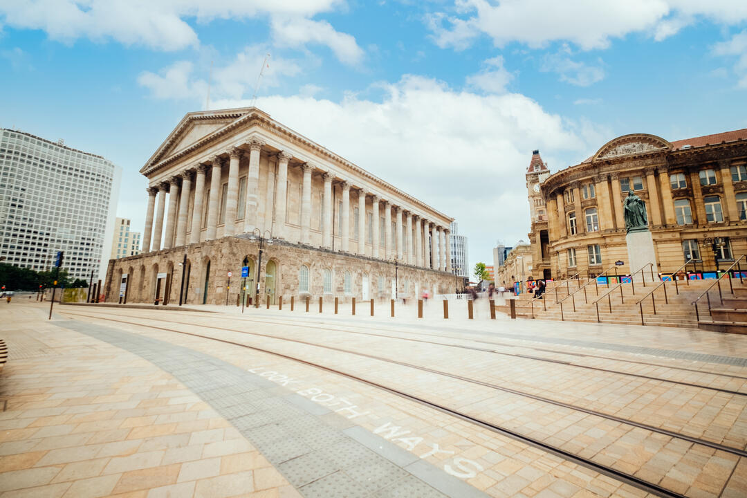 Wide plaza with historic stone buildings, including a neoclassical hall with columns and a curved civic building under a blue sky.