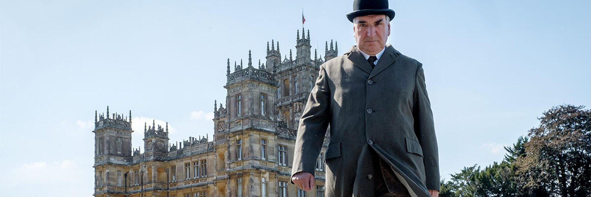 A person in period attire stands in front of a grand historic building with towers, set in landscaped gardens on a clear day.