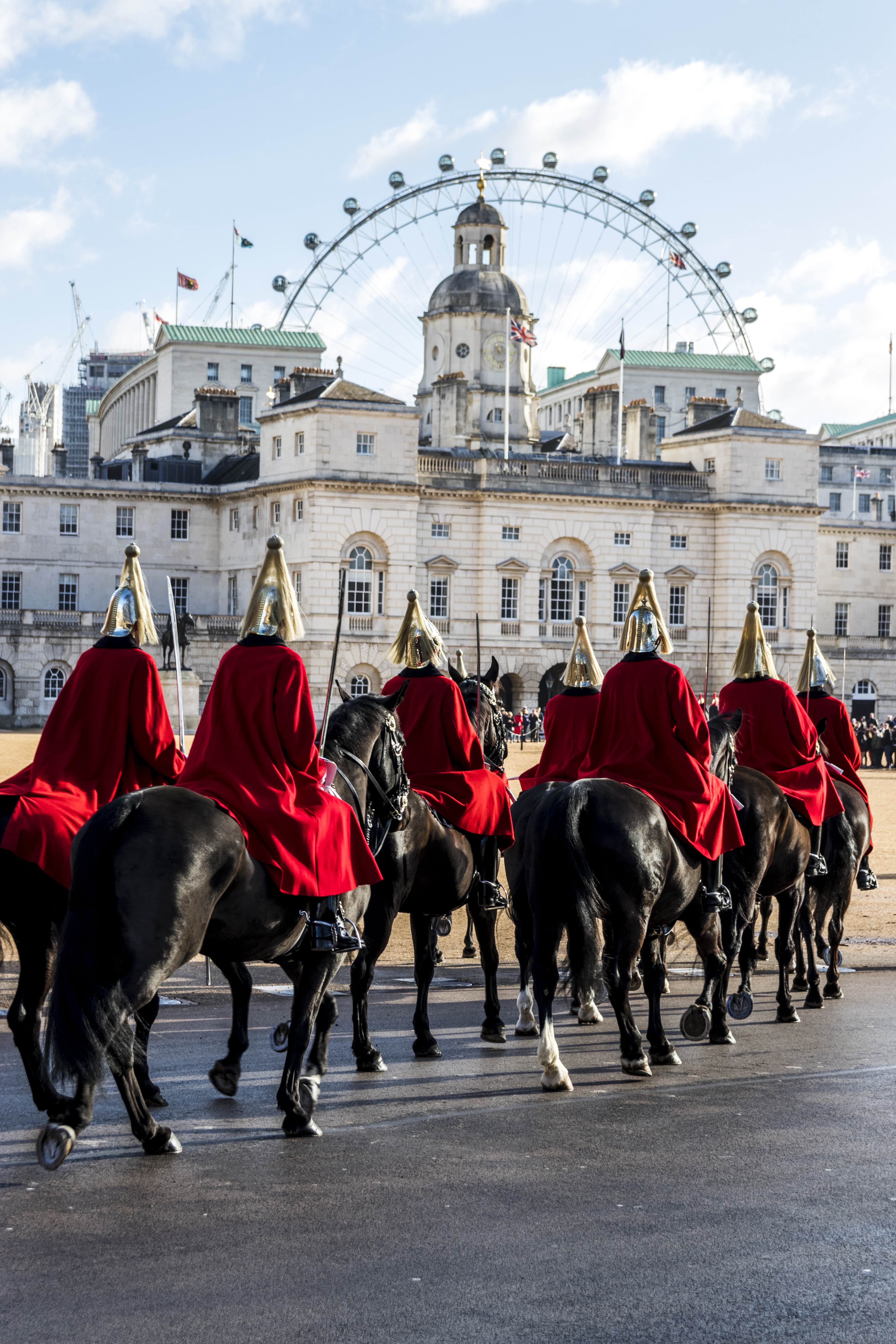 Men in ceremonial uniform riding horses towards a large historical building and a Ferris wheel