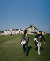 A man and a woman carrying golf equipment walking on the grounds of a country golf club.