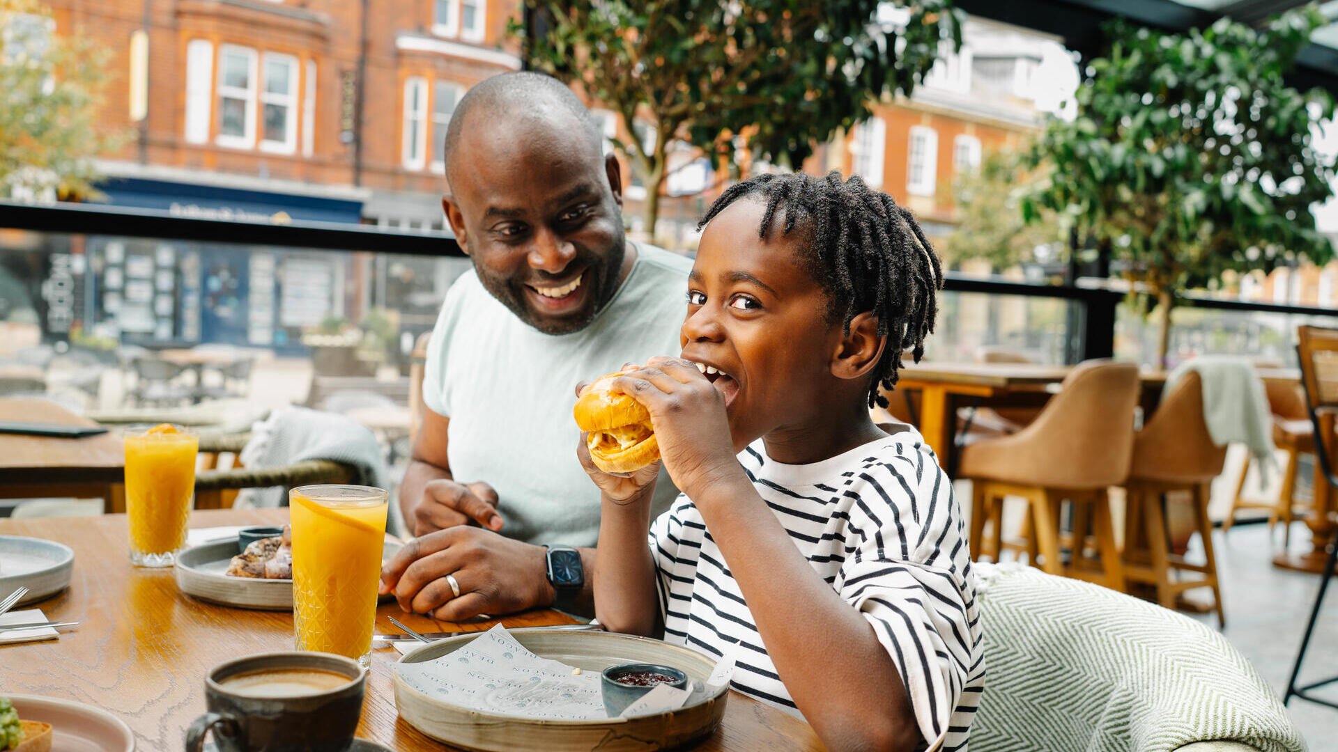 Child eating a burger and adult smiling at an outdoor restaurant table with drinks, plants, and red brick buildings in the background.