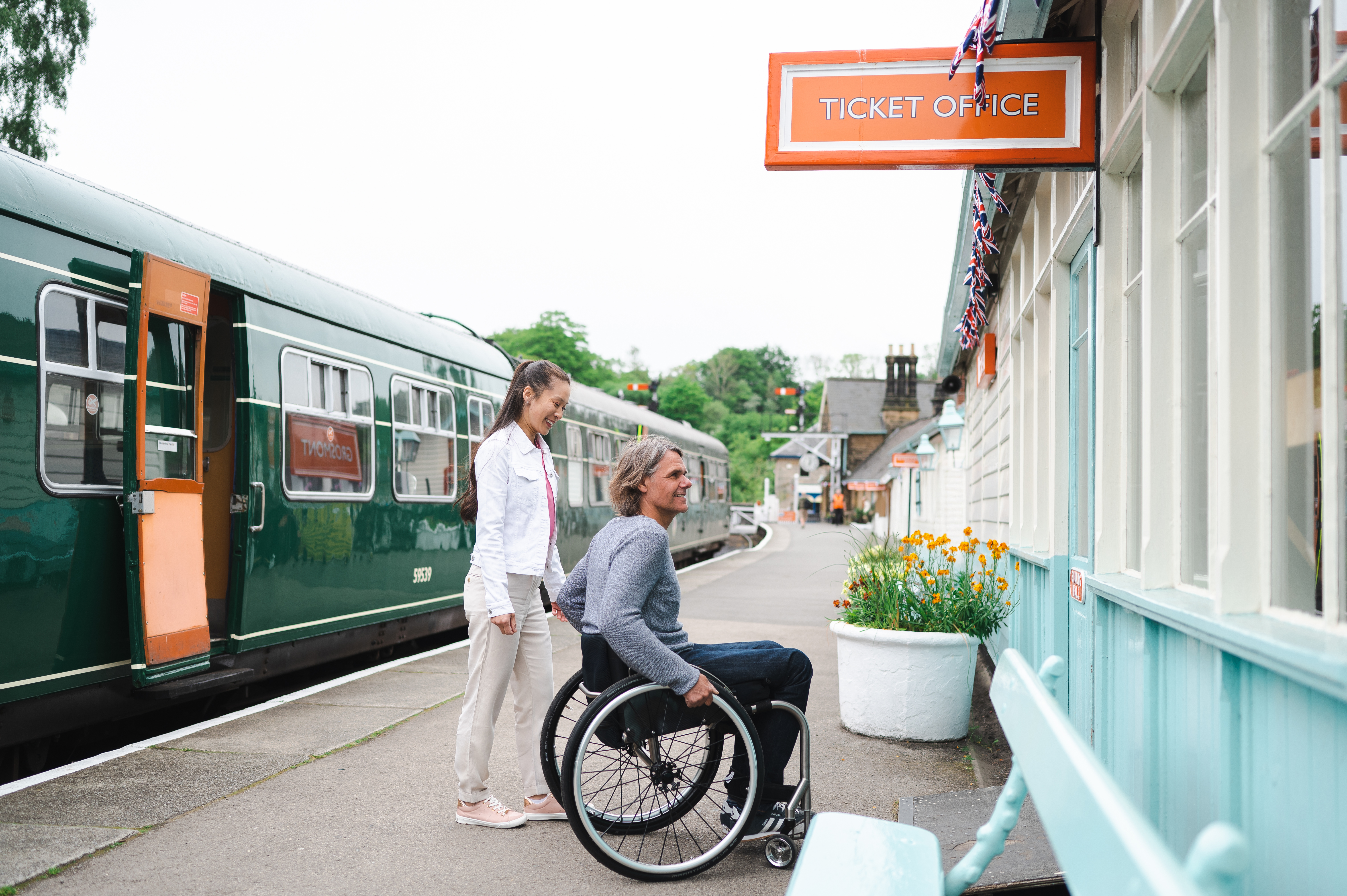 Man using a wheelchair and woman about to go into the ticket office at a station.