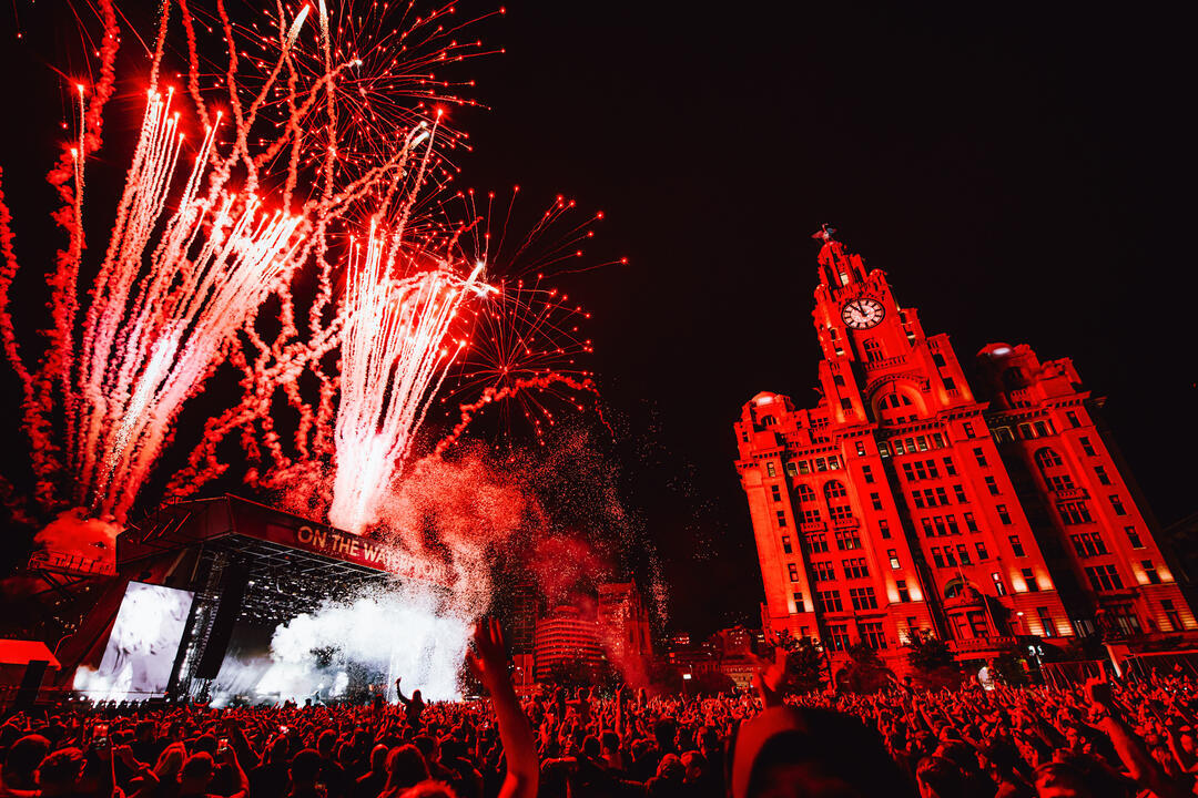Crowds of people watching a band at a music festival as fireworks explode, in front of the Liver Building.