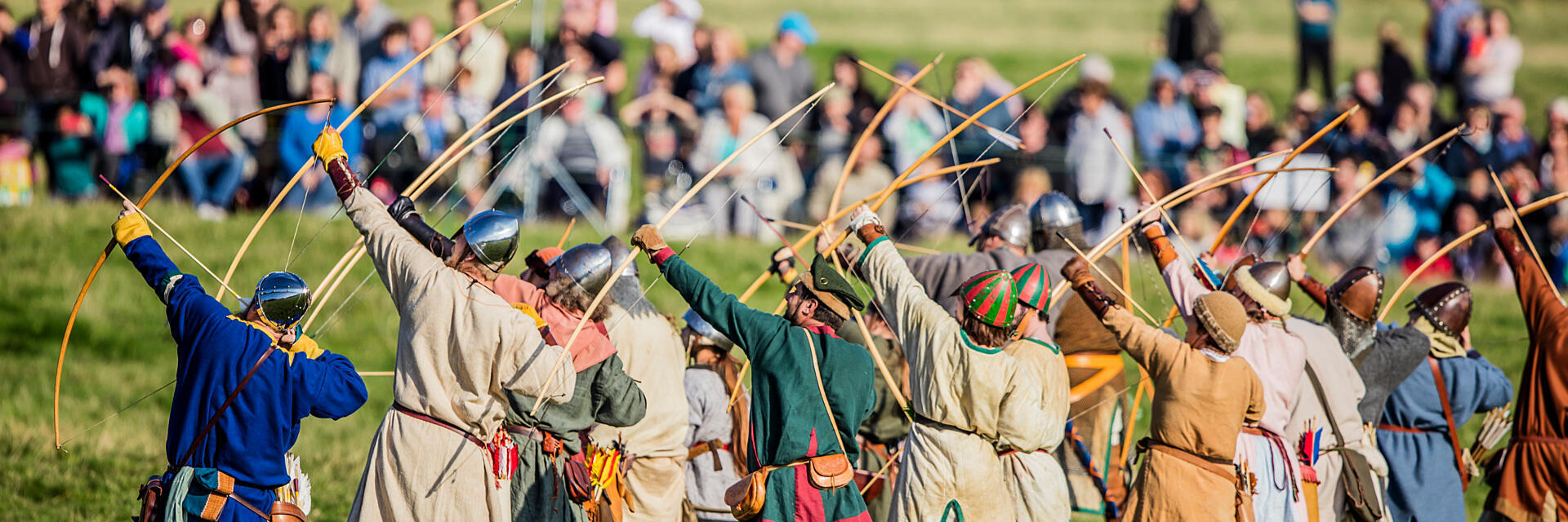 People in historical costumes reenact archery, drawing bows in a field with an audience in the background.