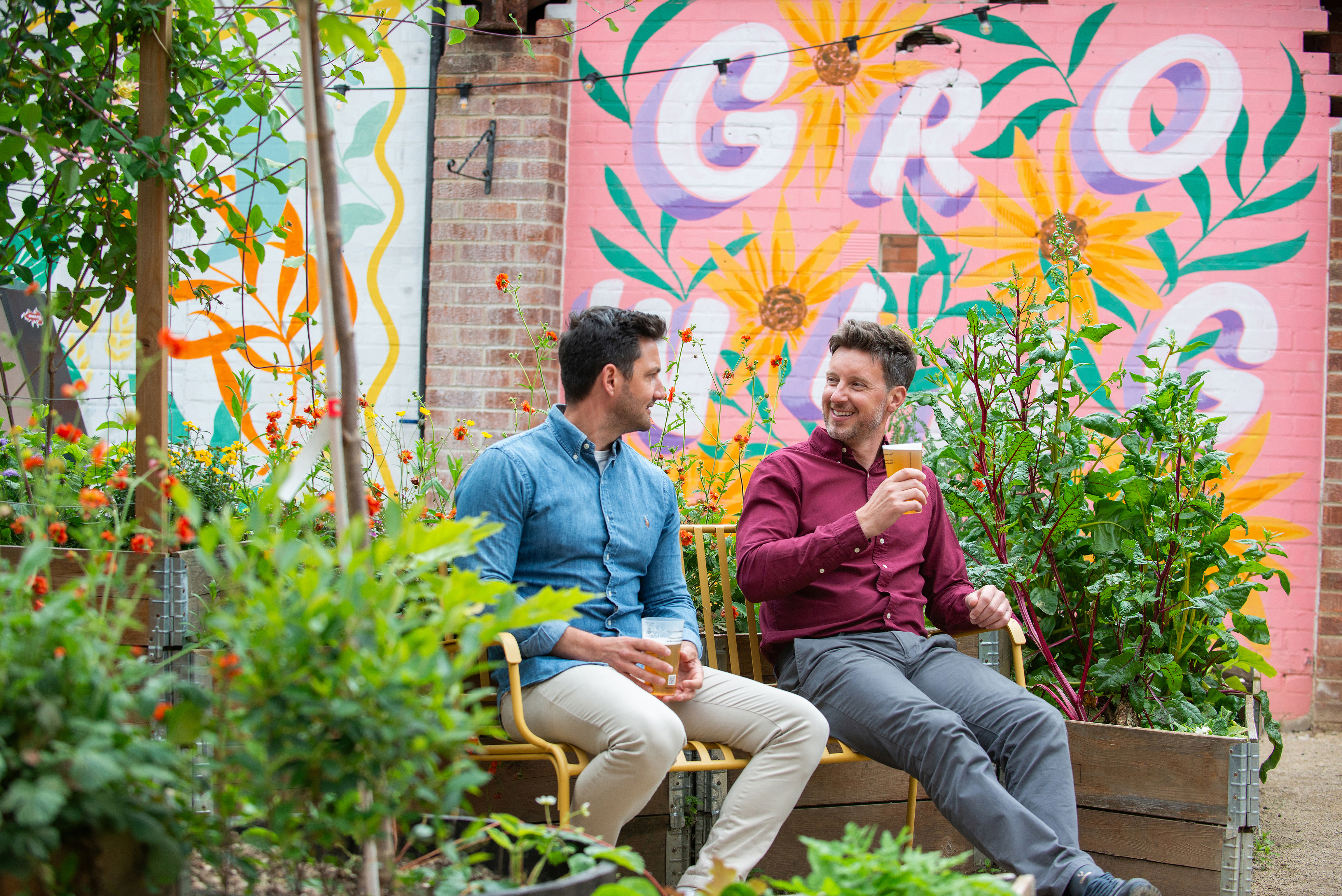 Two men sat on a bench in a garden with a drink