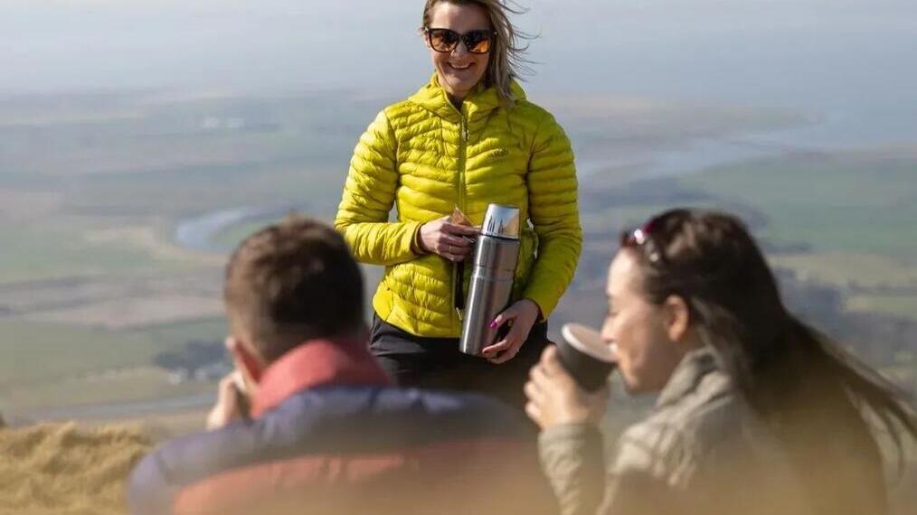 Three people enjoying a drink of coffee from a thermos flask on a hilltop