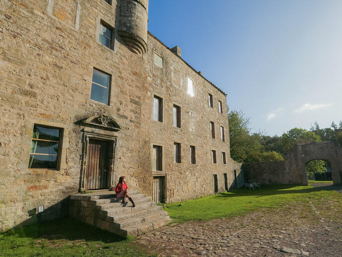 Historic stone building with arched entry gate, green lawn, and cobbled path. A person sits on steps in afternoon sunlight.