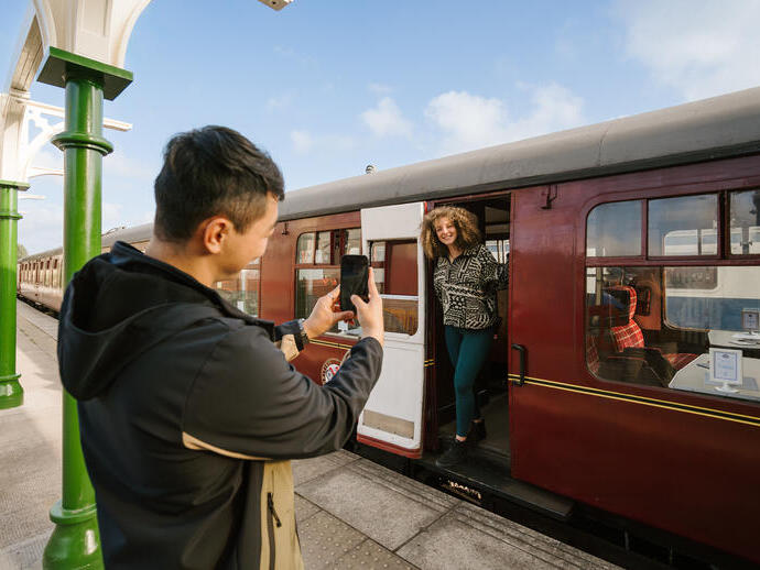 Un hombre tomando una foto de una mujer de pie junto a la puerta de un tren en el andén de una estación.
