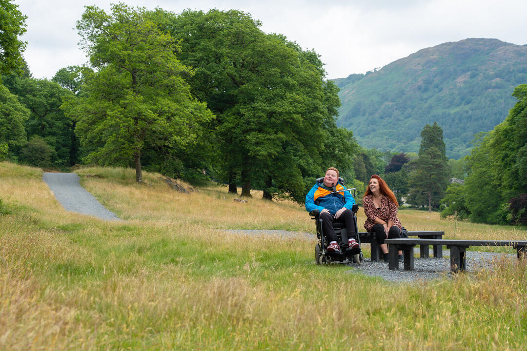 Two people enjoying a countryside view. One person is using an assisted wheelchair and the other person is sitting on a bench.