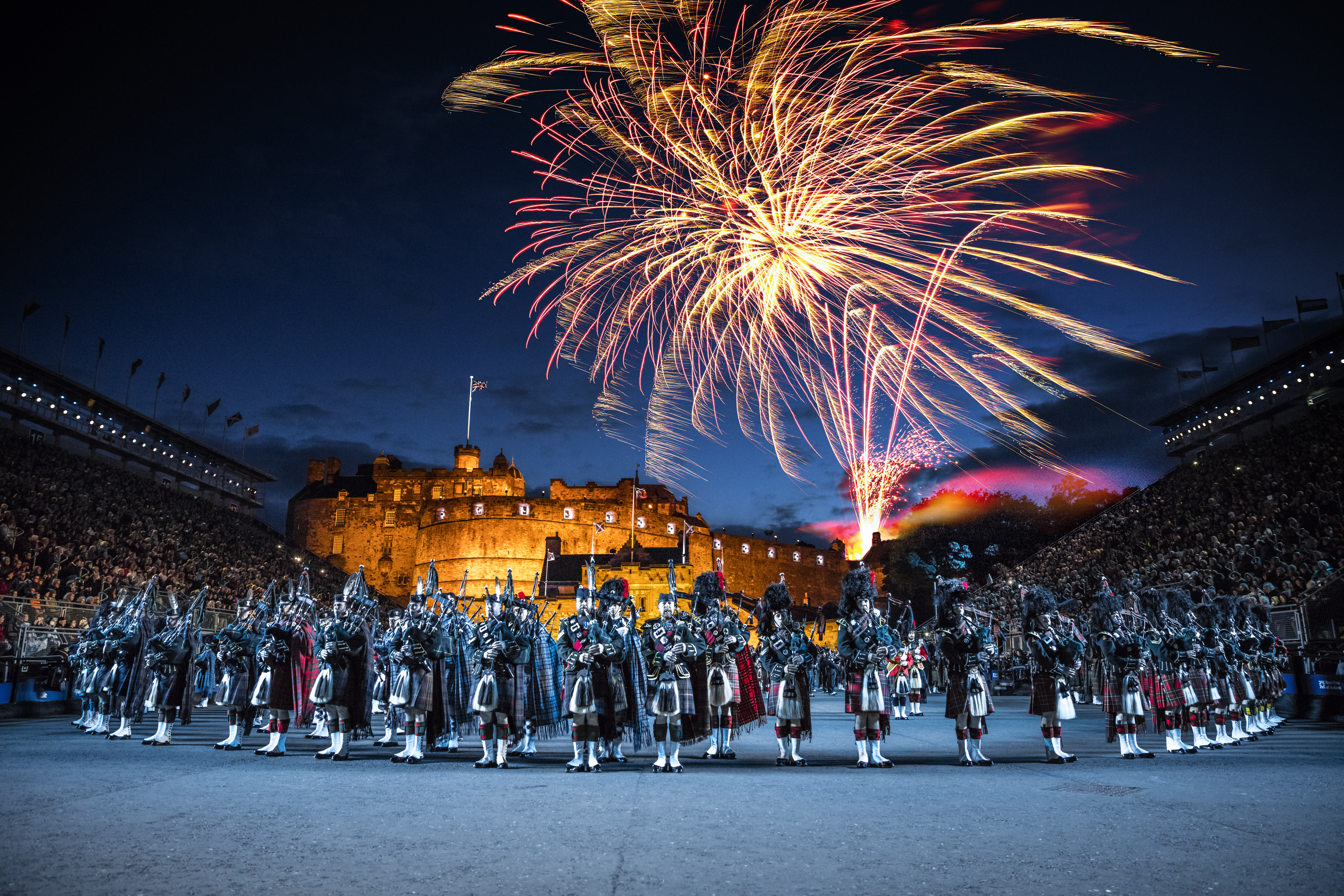La arena del Edinburgh Military Tattoo durante una actuación del evento militar, el patio de armas y las gradas repletas de espectadores. Un espectáculo de luces proyectado sobre las murallas del castillo. Banda de música con un director al frente y un grupo de gaiteros tocando la gaita. Fuegos artificiales explotando en el cielo nocturno.