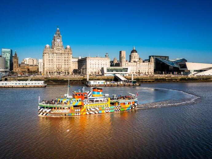 Colorful ferry on the River Mersey with Liverpool waterfront, historic buildings, and modern architecture under a clear blue sky.