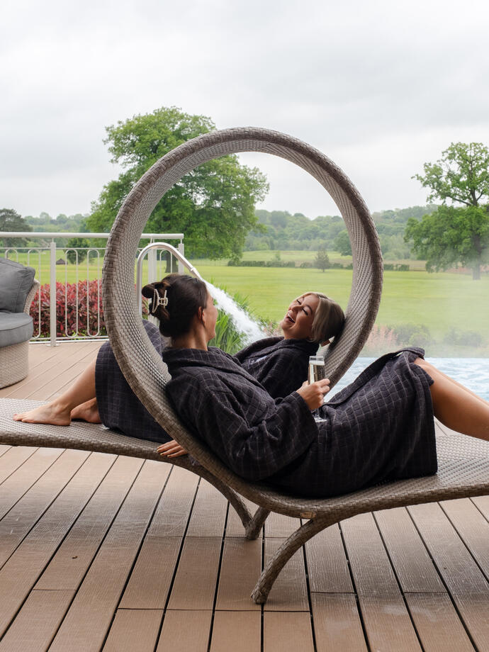Two women in robes lay on loungers with a garden beyond