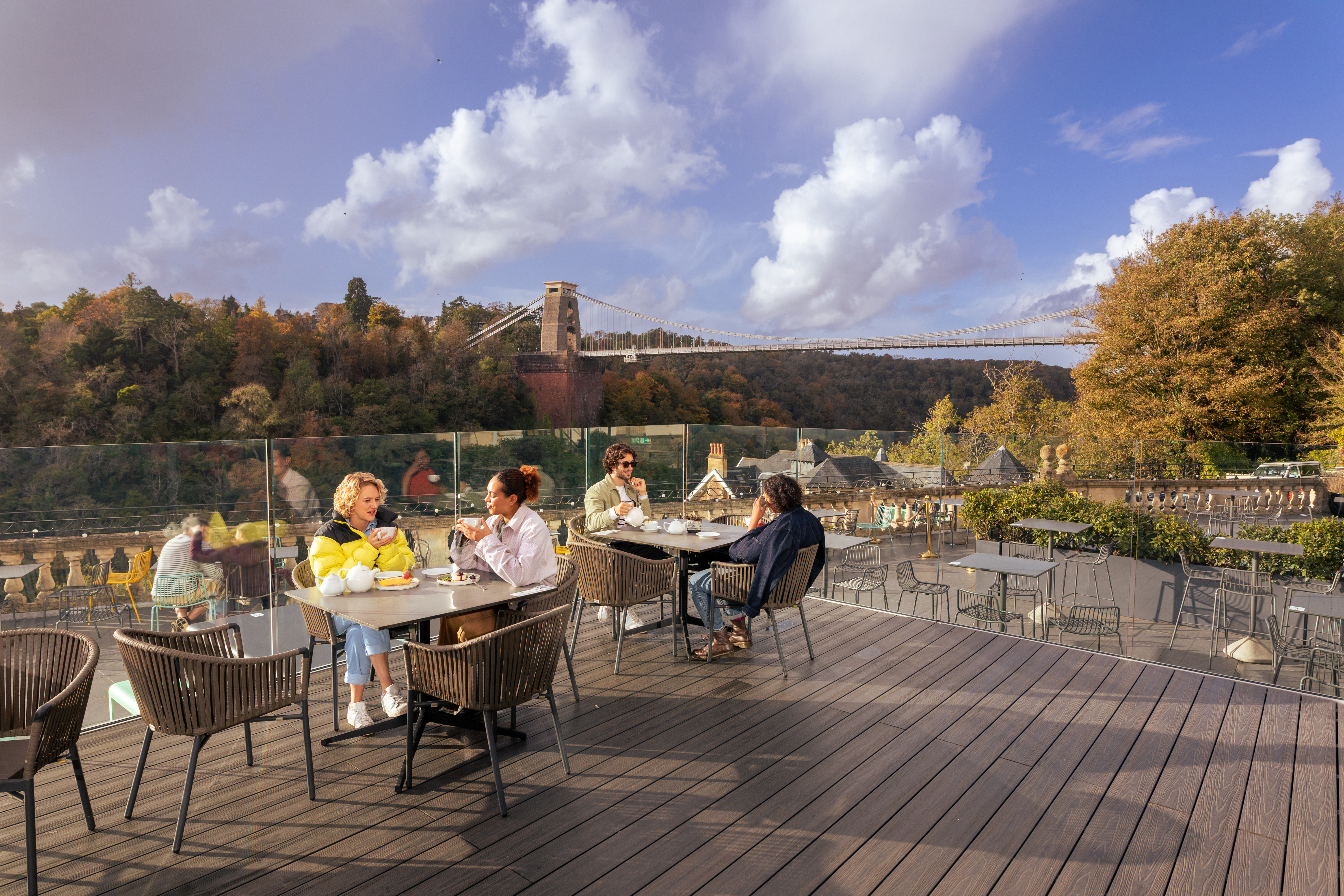 Friends enjoying tea and cake on a rooftop terrace, overlooking a river and suspension bridge.
