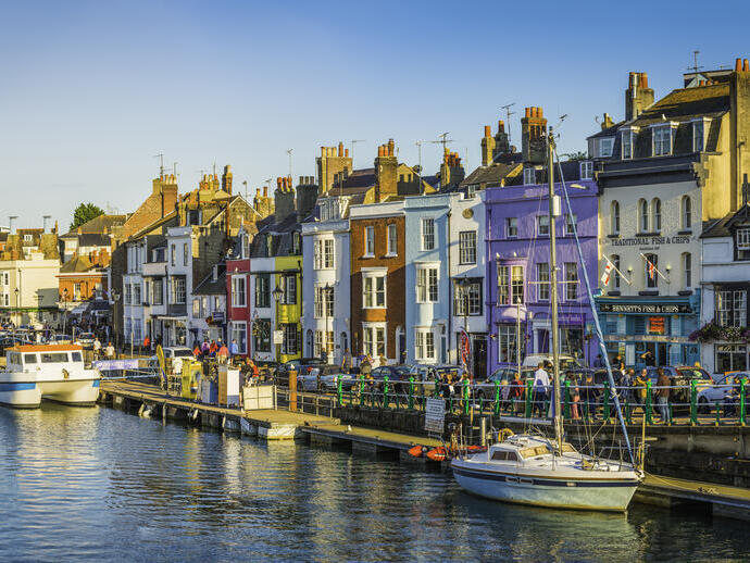People walking up and down the water front of a busy seaside town lined with colourful terraced houses and boats anchored in the harbour