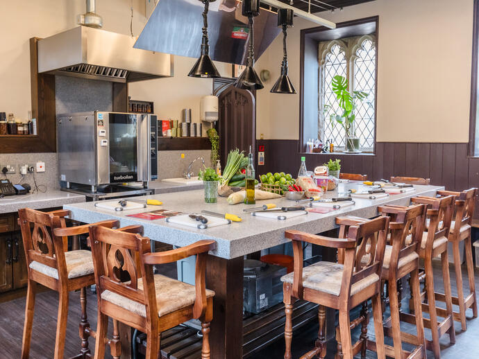 Modern kitchen with a central island, wooden chairs, prep stations, fresh ingredients, and a large oven. Bright window in the background.