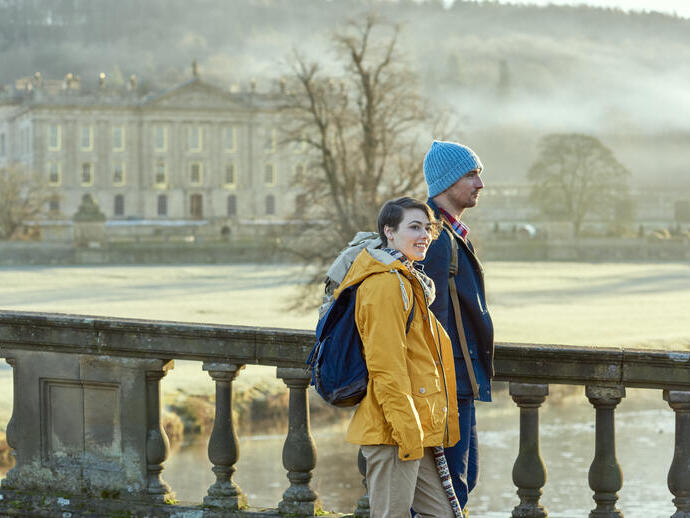 Pareja caminando por un puente de piedra, con una gran casa de campo al fondo