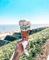 An ice cream cone being held in front of a beach, sea and beach huts