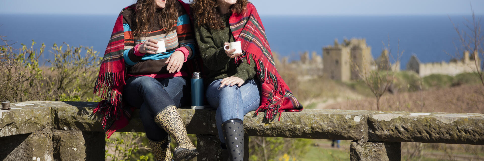 Two women with hot drinks, sitting on a stone wall near a castle