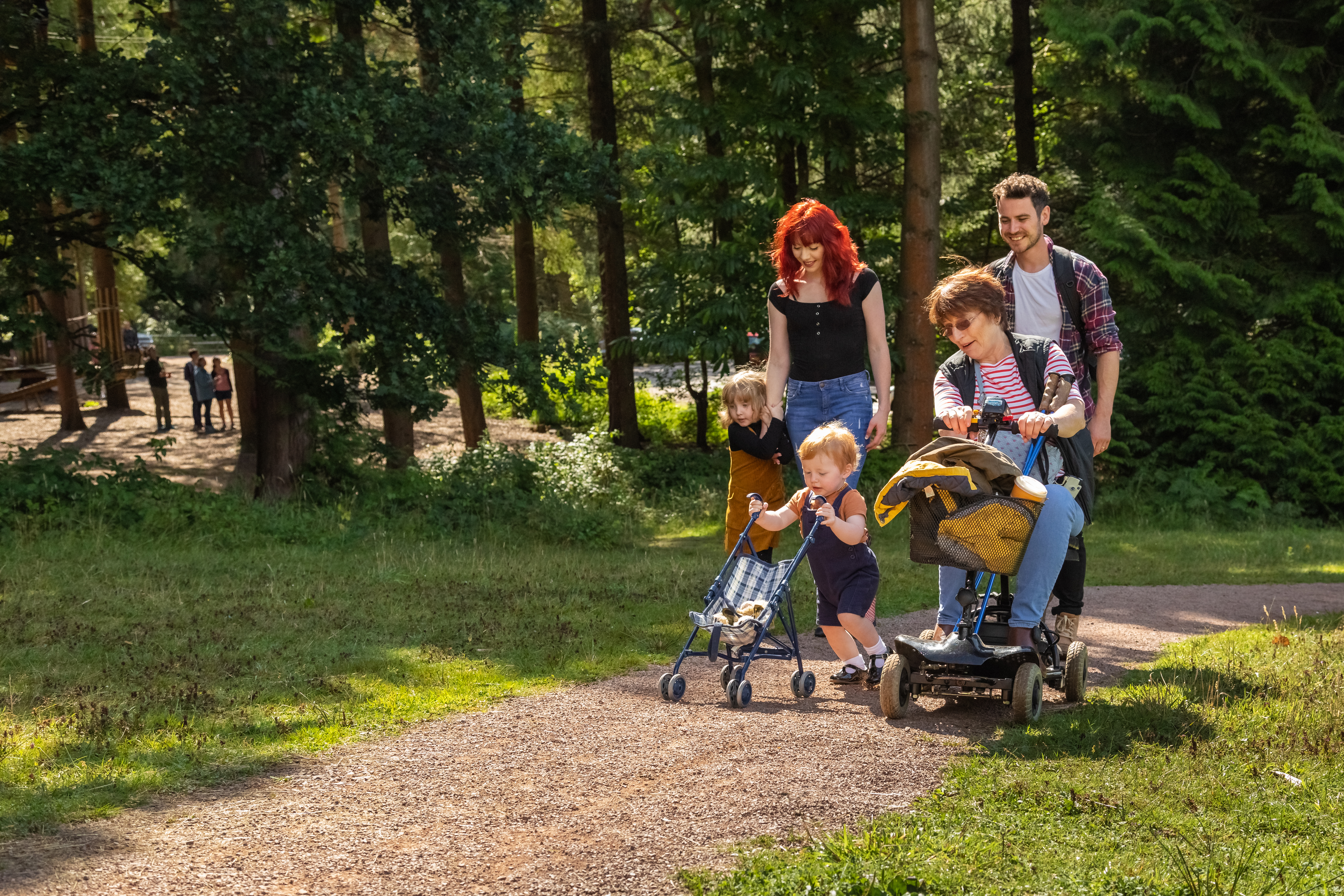A family enjoying a walk together in a forest, with children playing and an adult using a mobility scooter on the path.