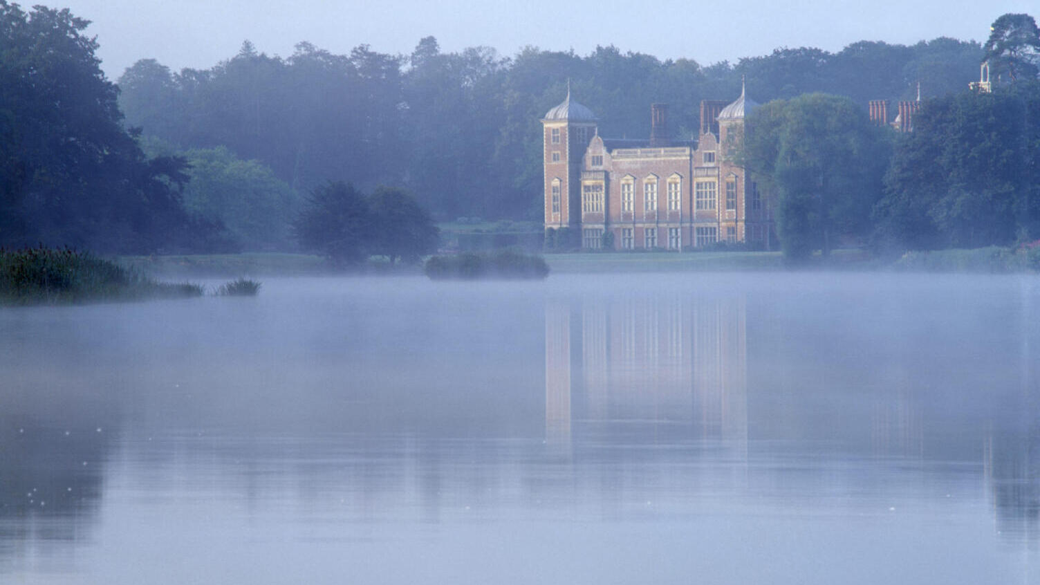 Early morning on the Blickling Hall estate with the house reflected in the water of the lake, and a bluish mist all around