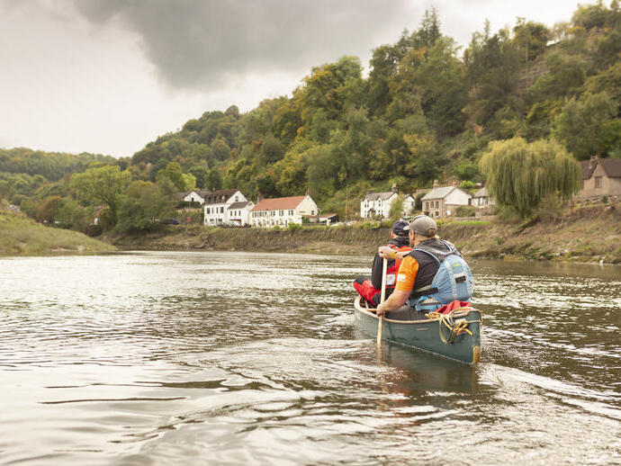 Two people canoe on a calm river with houses and forested hills under a cloudy sky in the background.