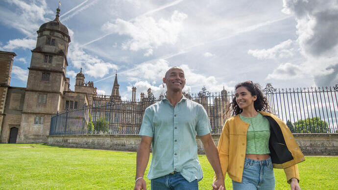 Two people holding hands, walking on grass outside a historic building with towers and iron fence under a partly cloudy sky.