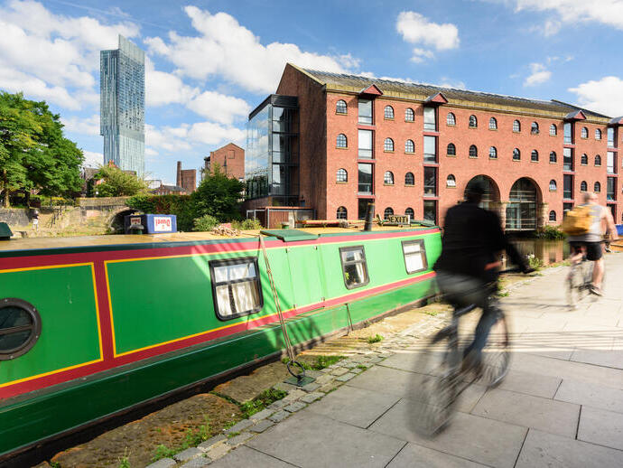 Canal boat moored by waterside, red brick warehouse, modern skyscraper, cyclists passing, sunny urban scene.
