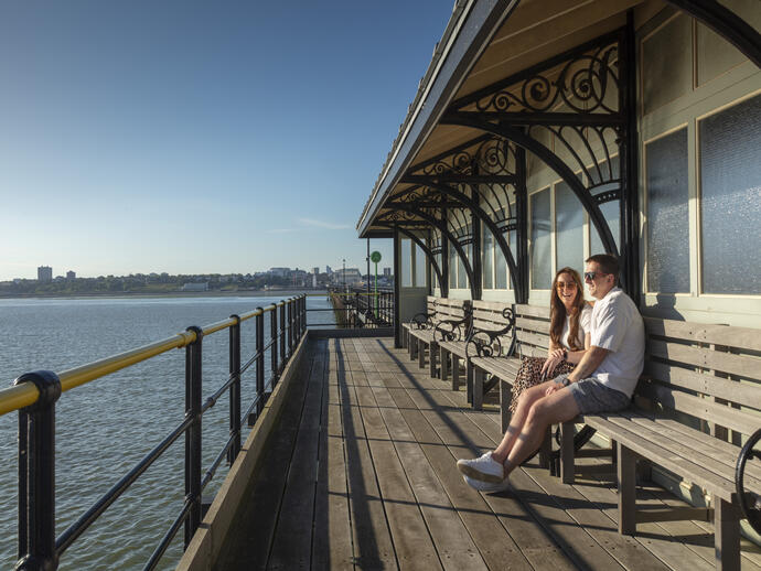 A man and a woman sit on a bench on a pier at the beach
