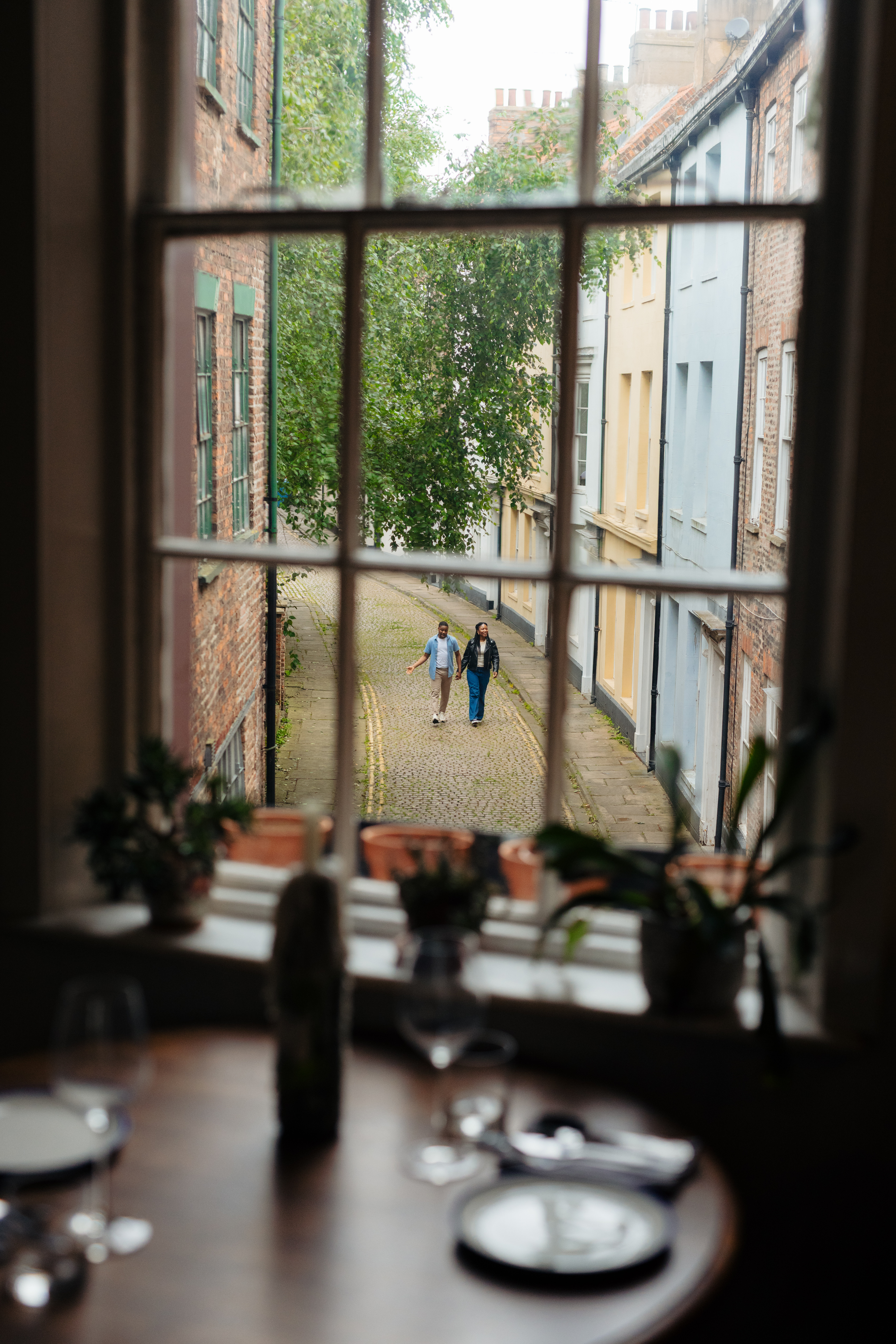 Vista desde una ventana a una calle adoquinada con dos personas caminando y una mesa de comedor desenfocada en primer plano.
