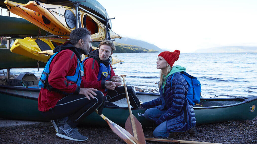 A group of people on the shore of a lake next to a rack of kayaks and canoes