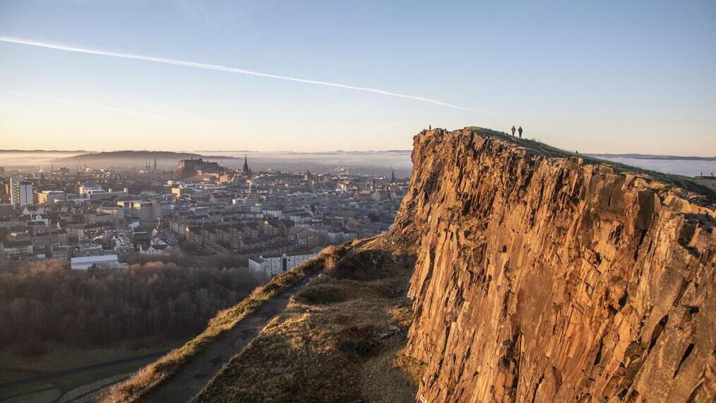 Panoramablick auf eine Stadt am Fuße einer Felsklippe unter klarem Himmel.