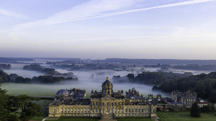 Grande maison de campagne vue d'en haut avec des jardins à la française et une campagne boisée derrière, recouverte de nuages bas