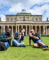 Two men and a woman sit on grass outside of a heritage building