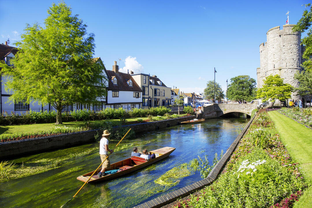 A man and a couple in a punt on a small river surrounded by gardens and houses