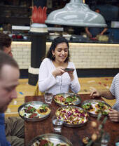 Woman sat with friends at a table, taking a photo of food in a food court
