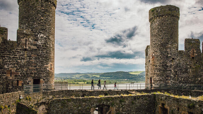 Stone castle ruins with two large round towers, people walking on a wall, hills and water visible in the background under a cloudy sky.