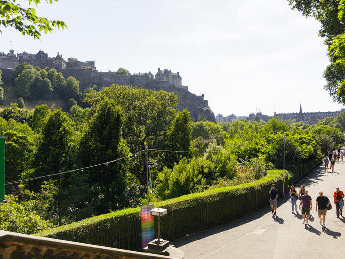 Vista del castillo de Edimburgo rodeado de árboles, con personas caminando por un sendero soleado en primer plano.