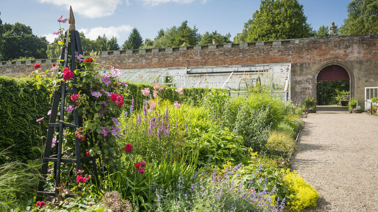 Lush garden with colorful flowers, a wooden trellis, gravel path, brick wall, and glasshouse under a blue sky with scattered clouds.