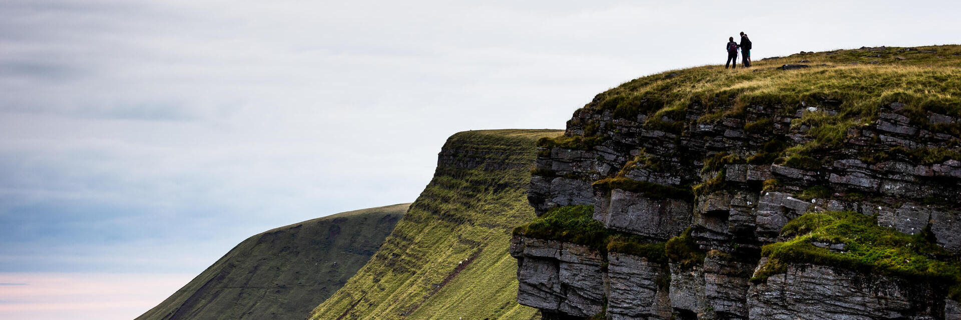 Two people stand atop a grassy cliff with dramatic rocky edges, overlooking rolling green hills under a cloudy sky.